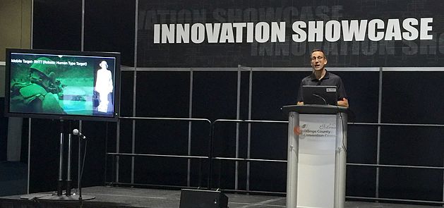 Presenter at the I/ITSEC Innovation Showcase podium, with a screen displaying the RhTT mobile target system.