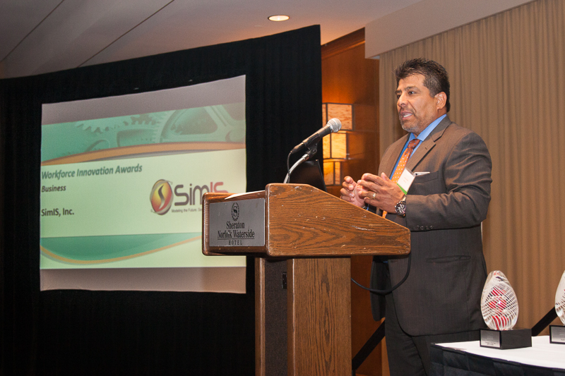 Dr. Johnny Garcia speaking at a podium, with a screen behind him reading 'Workforce Innovation Awards, Business, SimIS, Inc.'
