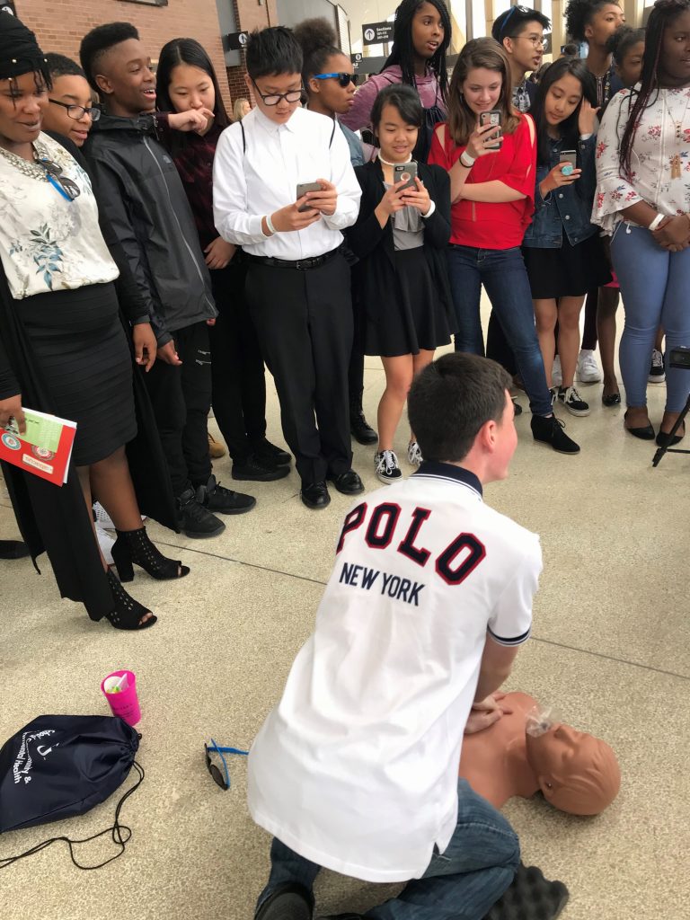 A student practices CPR on a training mannequin using the AIMS system while a group of students watches and records on their phones.