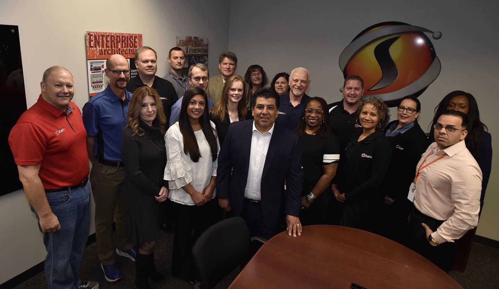 Dr. Johnny Garcia with the SimIS team gathered in the office conference room, SimIS logo on the wall.