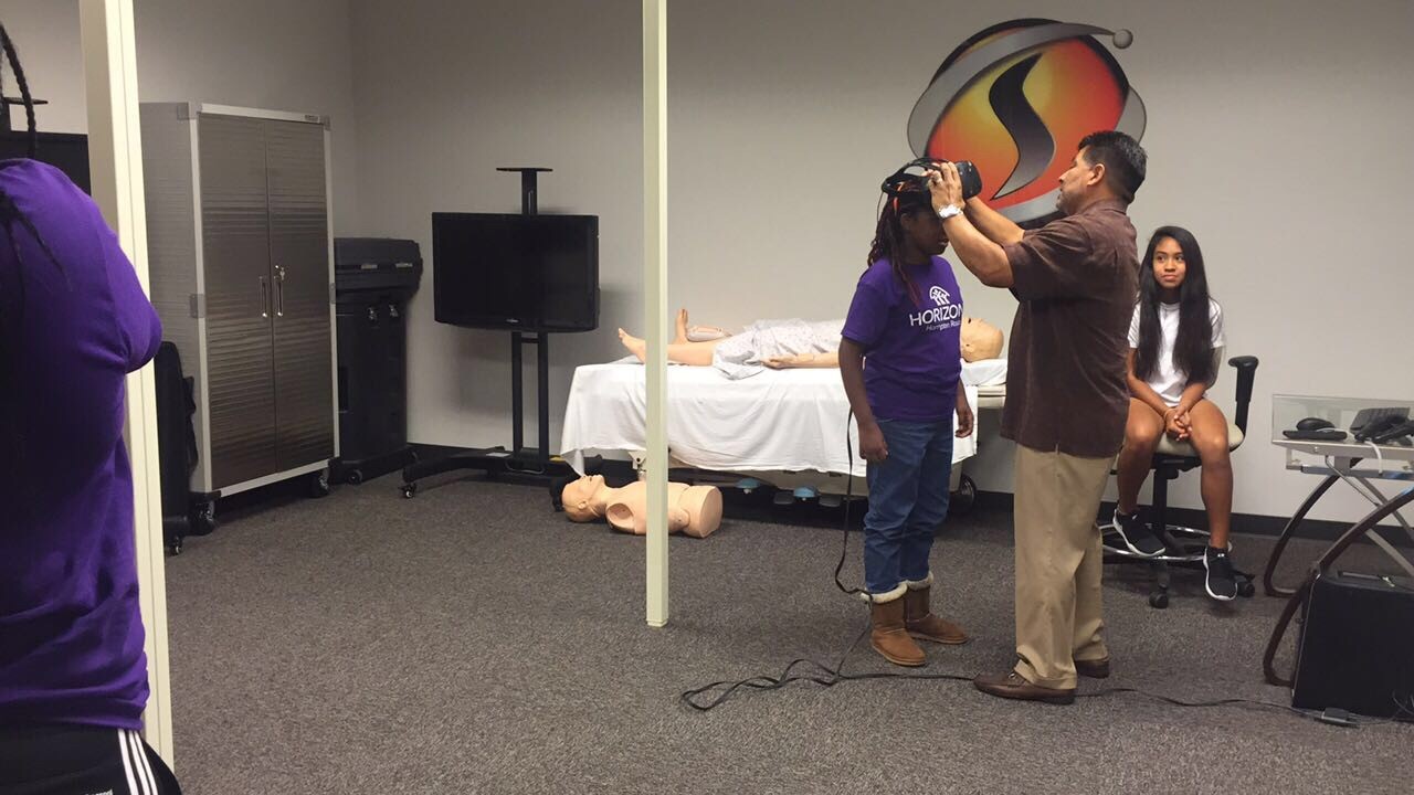 Alt text: "A SimISDr. Garcia helps a Horizons camper put on a VR headset, with medical training mannequins in the background.