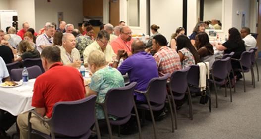 Attendees seated at long tables enjoying lunch during the JT&E-Suffolk summer cookout.