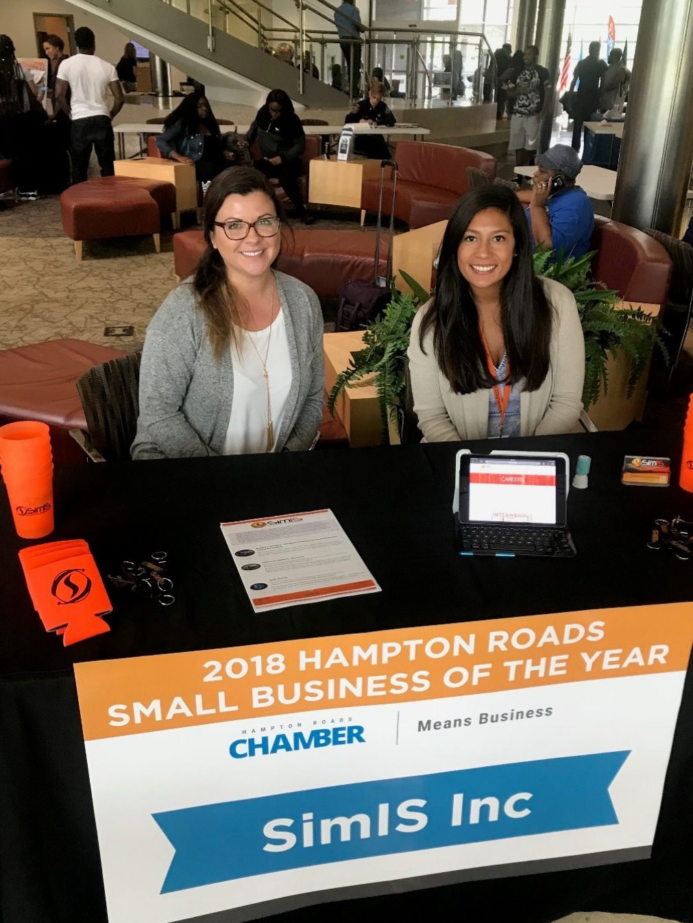 Two SimIS representatives seated at an information table displaying the 2018 Hampton Roads Small Business of the Year banner at TCC's Campus Takeover.
