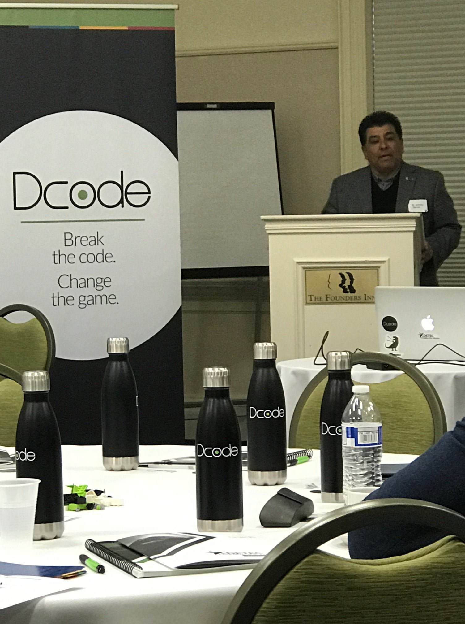 Dr. Johnny Garcia speaking at a podium at the Dcode event, with branded water bottles on the table and a Dcode banner reading 'Break the code. Change the game.'