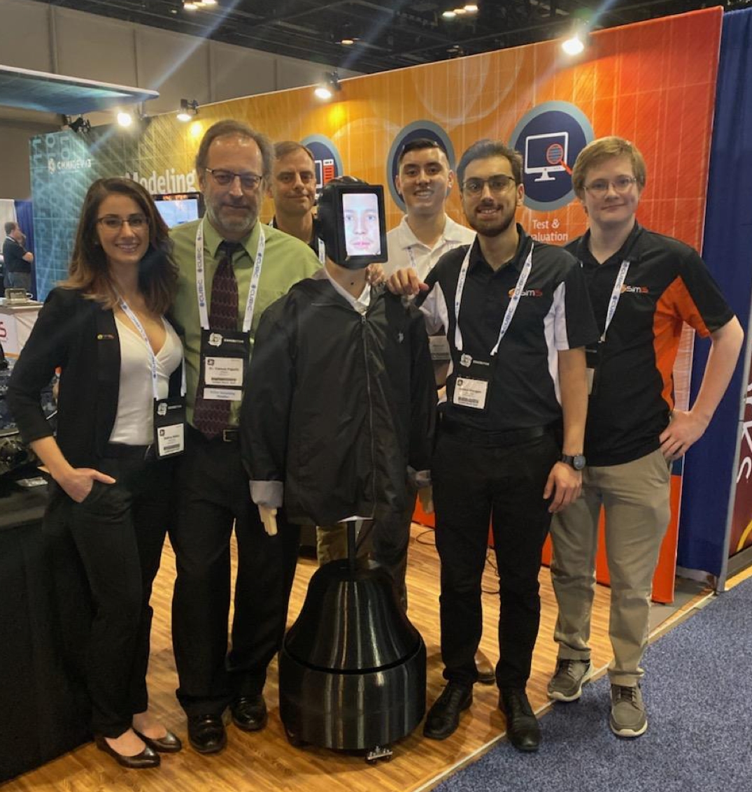 SimIS team members posing with the ADRT robotic trainer, featuring a digital face display, at the IITSEC booth.