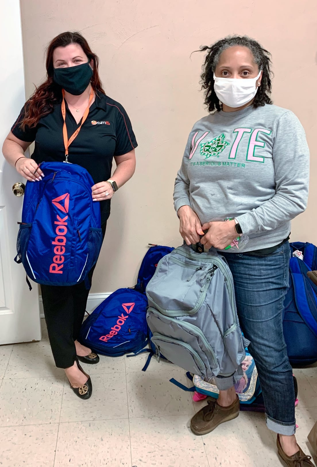A masked SimIS employee and a Wesley Community Service Center representative holding donated backpacks.