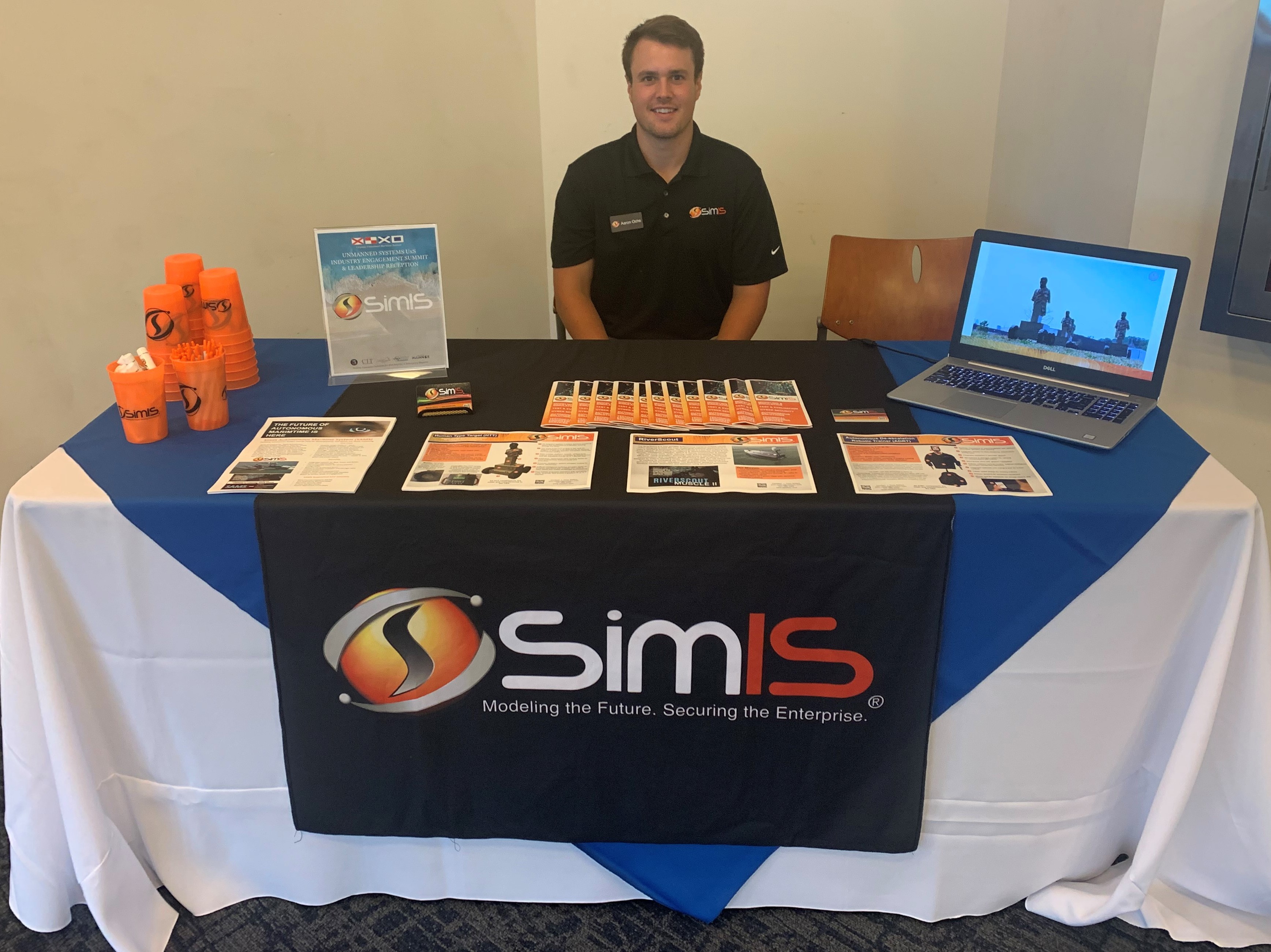 Alt text for Image 1: "SimIS representative at the company's booth, with product handouts for HTT, RiverScout, and ADRT displayed on the table.