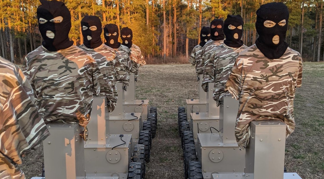 Two rows of camouflage-clad robotic training targets with masked human-like heads positioned along an outdoor wooded training range.